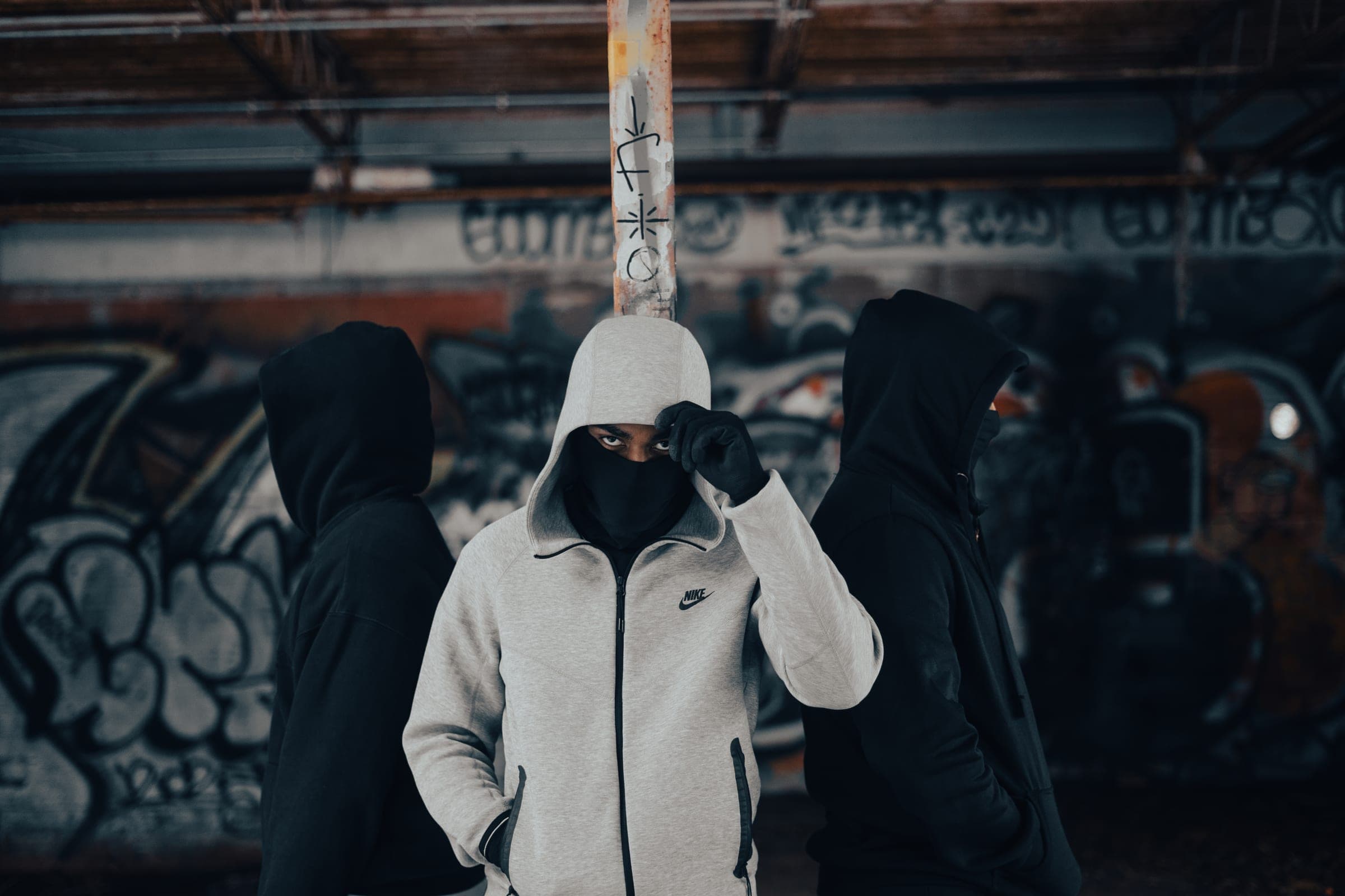 Three masked figures in hoodies against a graffiti wall